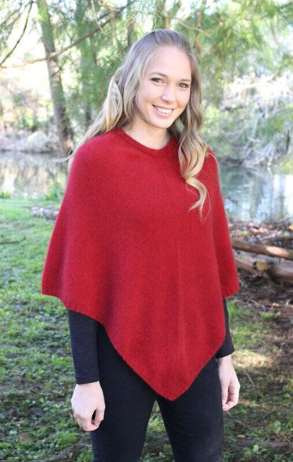 Red possum and merino wool poncho on female model standing in garden. Warm, soft, lightweight possum and merino wool. Lothlorian